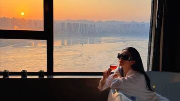 A person sitting by a window with a view of a city skyline and a body of water during sunset.