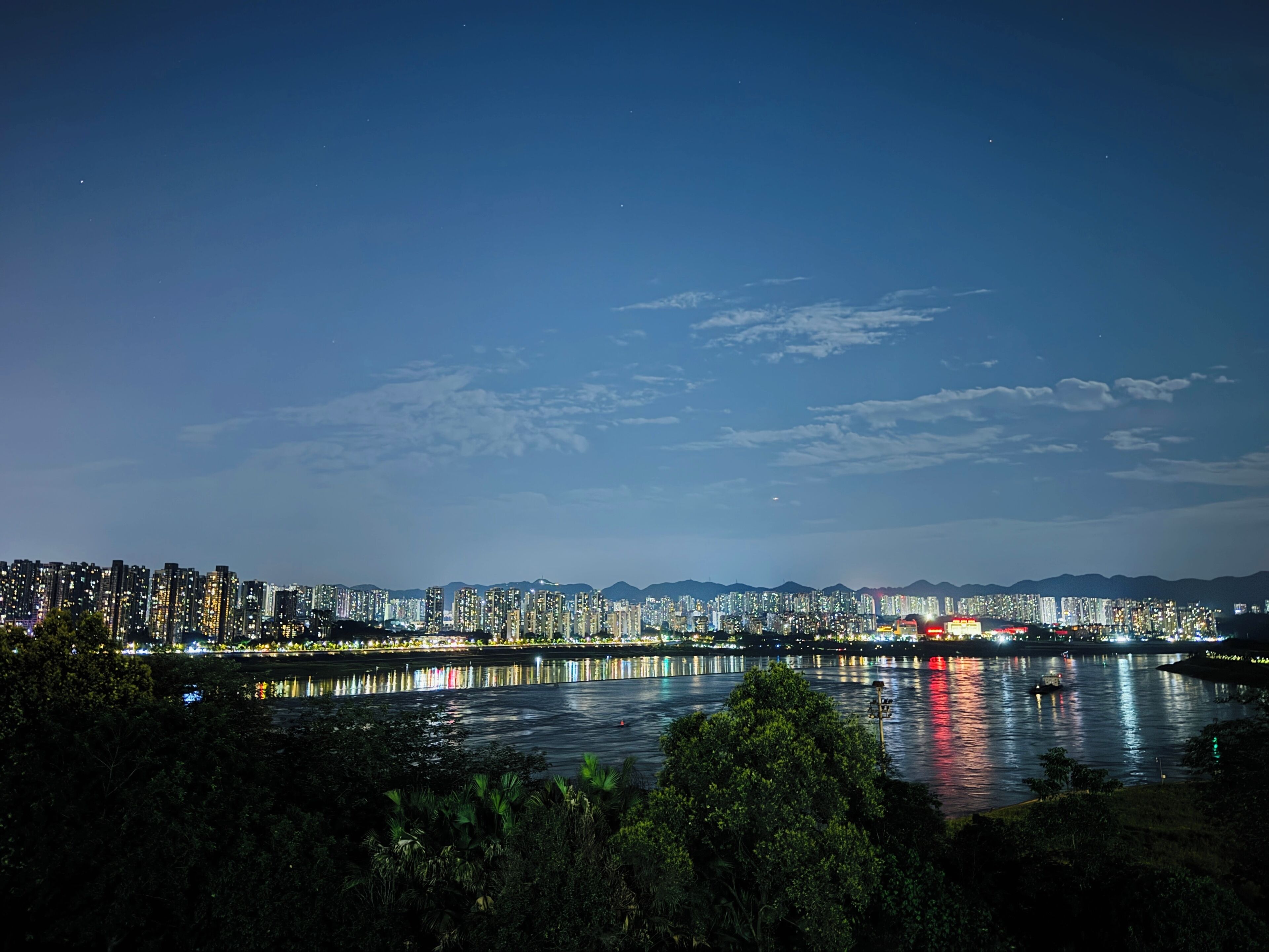 A nighttime cityscape with a river reflecting lights, high-rise buildings, and mountains in the background.
