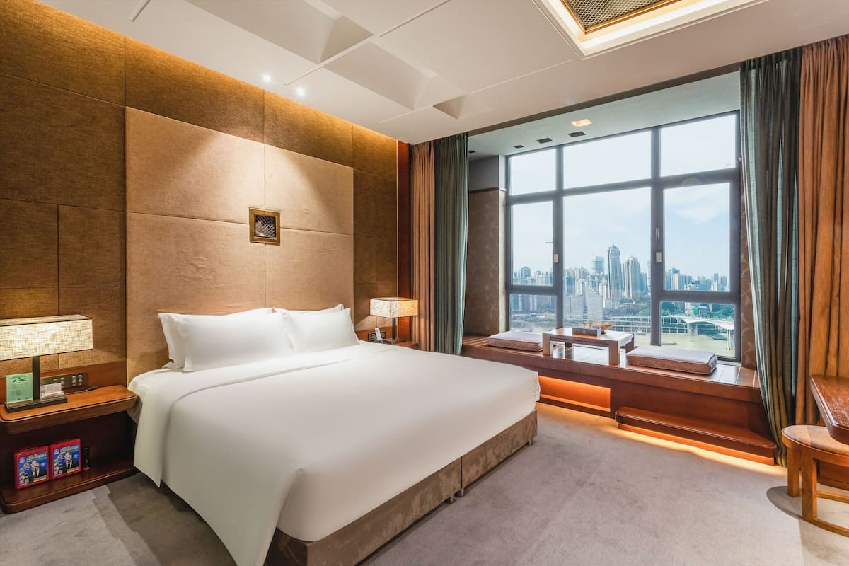 A hotel room with a large bed, a nightstand with a lamp, a view of the city through large windows, and a seating area by the window.