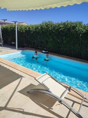A swimming pool with two people in the water, surrounded by a tiled deck and a covered area.