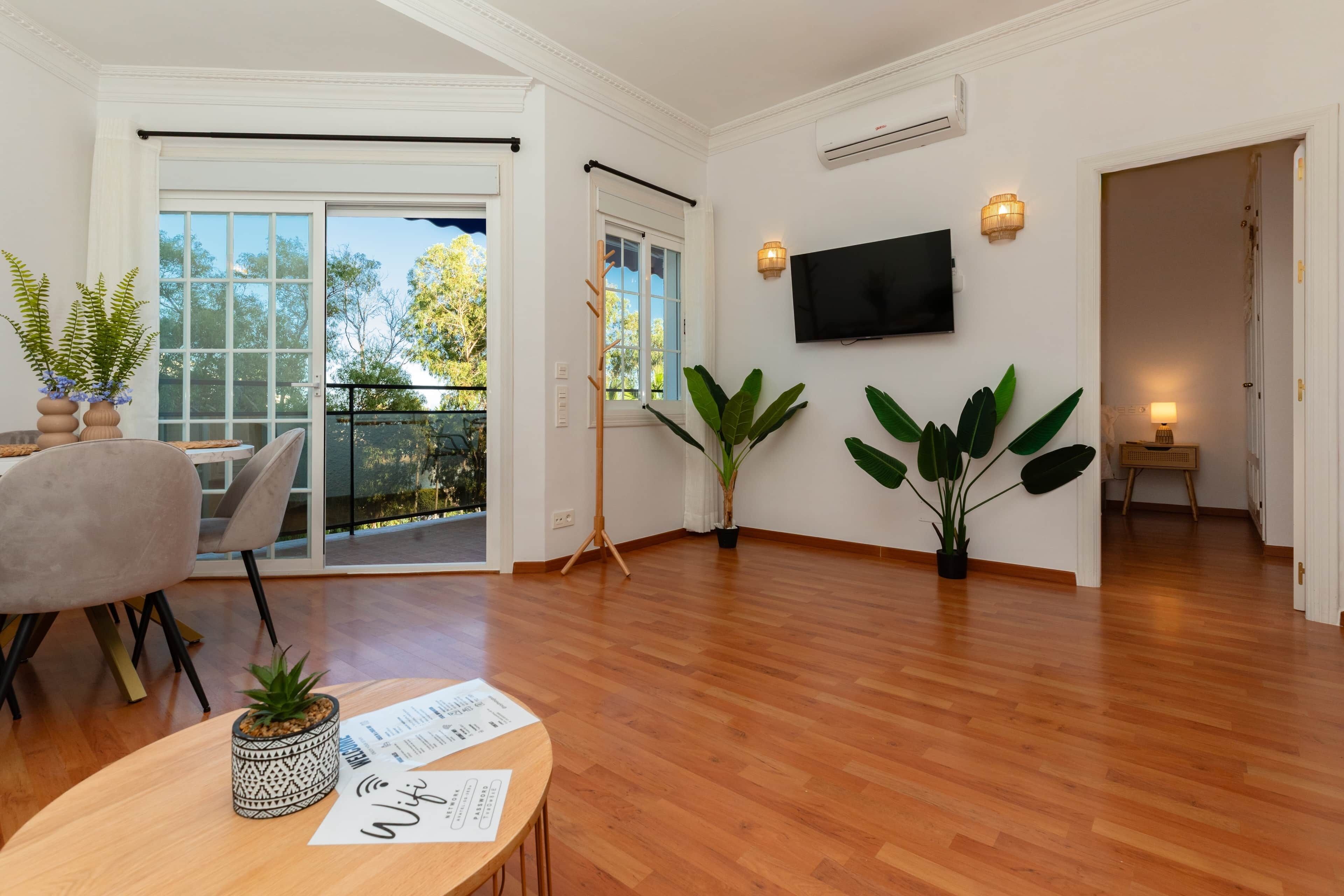 A spacious living room with a wooden floor, a dining area with a table and chairs, and a balcony with a view of trees.