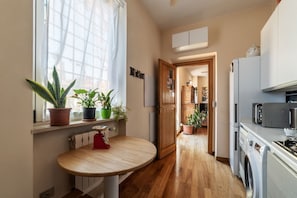A narrow kitchen with wooden flooring, a built-in oven, a microwave, and a washing machine. There are potted plants on the windowsill and a small table with a red chair.