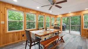 Une pièce spacieuse avec une grande table en bois, plusieurs chaises et un banc. De grandes fenêtres offrent une vue sur la verdure environnante.