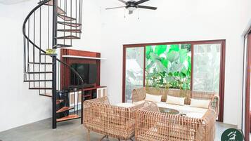 A modern living room with a spiral staircase, a flat-screen TV, wicker furniture, and a large window with a view of greenery.