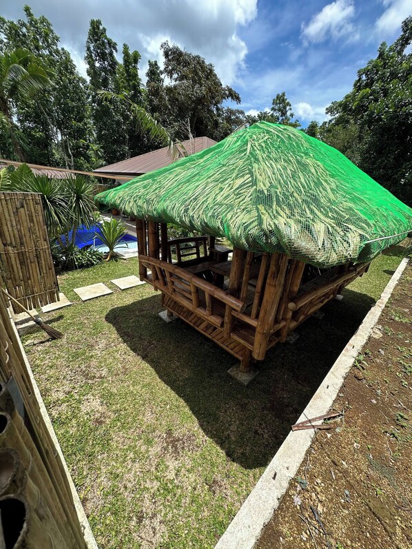 A wooden hut with a thatched roof, surrounded by a bamboo fence, and a pool in the background.