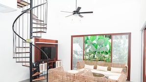 A modern living room with a spiral staircase, a ceiling fan, a large window with a view of greenery, and a wicker sofa set.