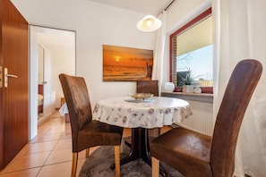 Un espace repas comprenant une table, des chaises, un tableau représentant un coucher de soleil, une fenêtre avec des rideaux et une vue sur l’extérieur.