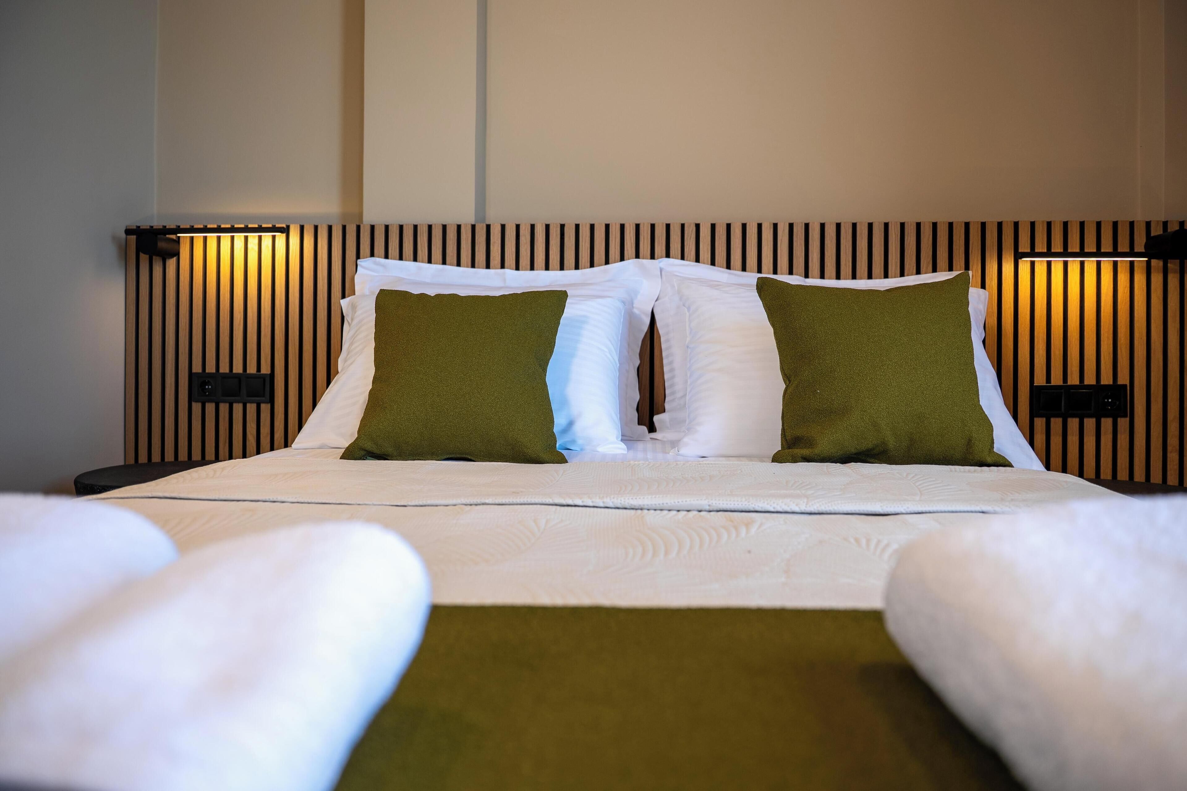 A neatly made bed with white linens and two green pillows, a wooden slat headboard, and a bedside table with a lamp.