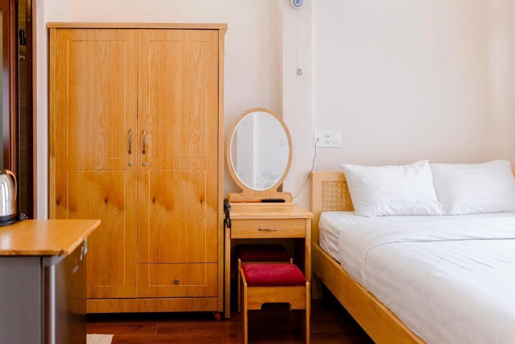 A bedroom with a wooden wardrobe, a bedside table with a mirror, a bed with white bedding, and a red bench.