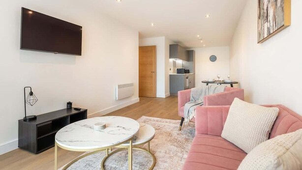 A modern living room with a flat-screen TV, a marble coffee table, a pink sofa, and a small kitchen area in the background. - Spacious 1BR Apartment in Central Leeds (Leeds)