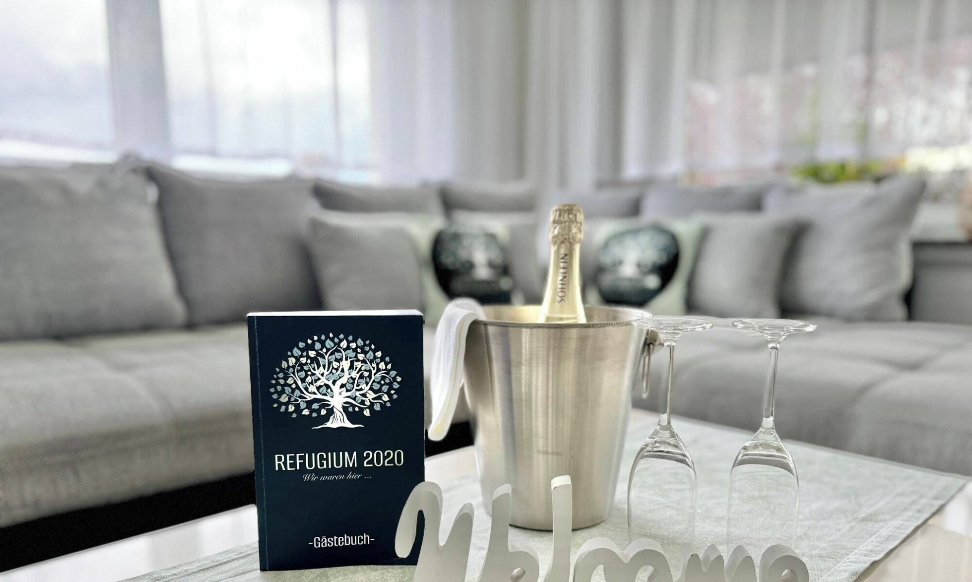 A book titled 'Refugium 2020' on a table, a champagne bottle in a silver bucket, and two wine glasses.