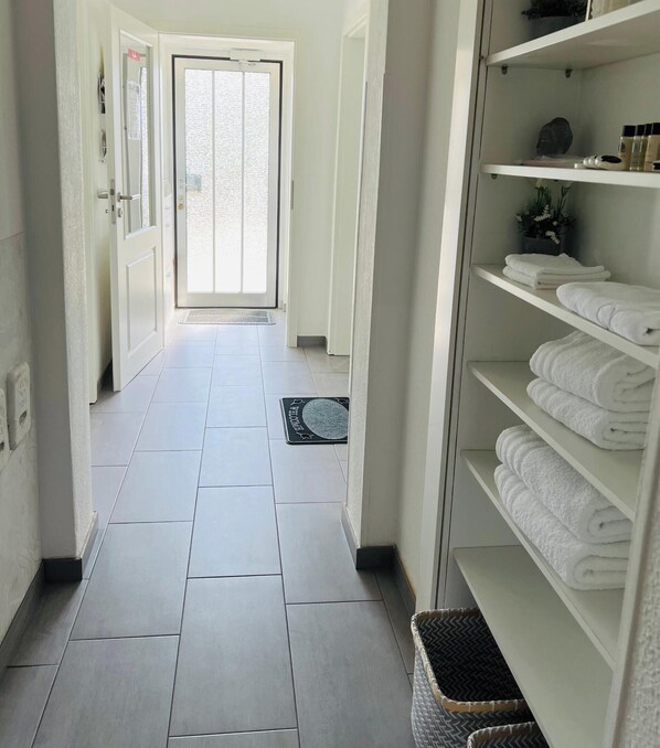 A hallway with a tiled floor, a door leading to a bright room, and a shelf with neatly folded towels. - Holiday Home '2 Apartments in Refugium 2020' with Private Terrace, Private Garden and Wi-Fi (Marienheide)