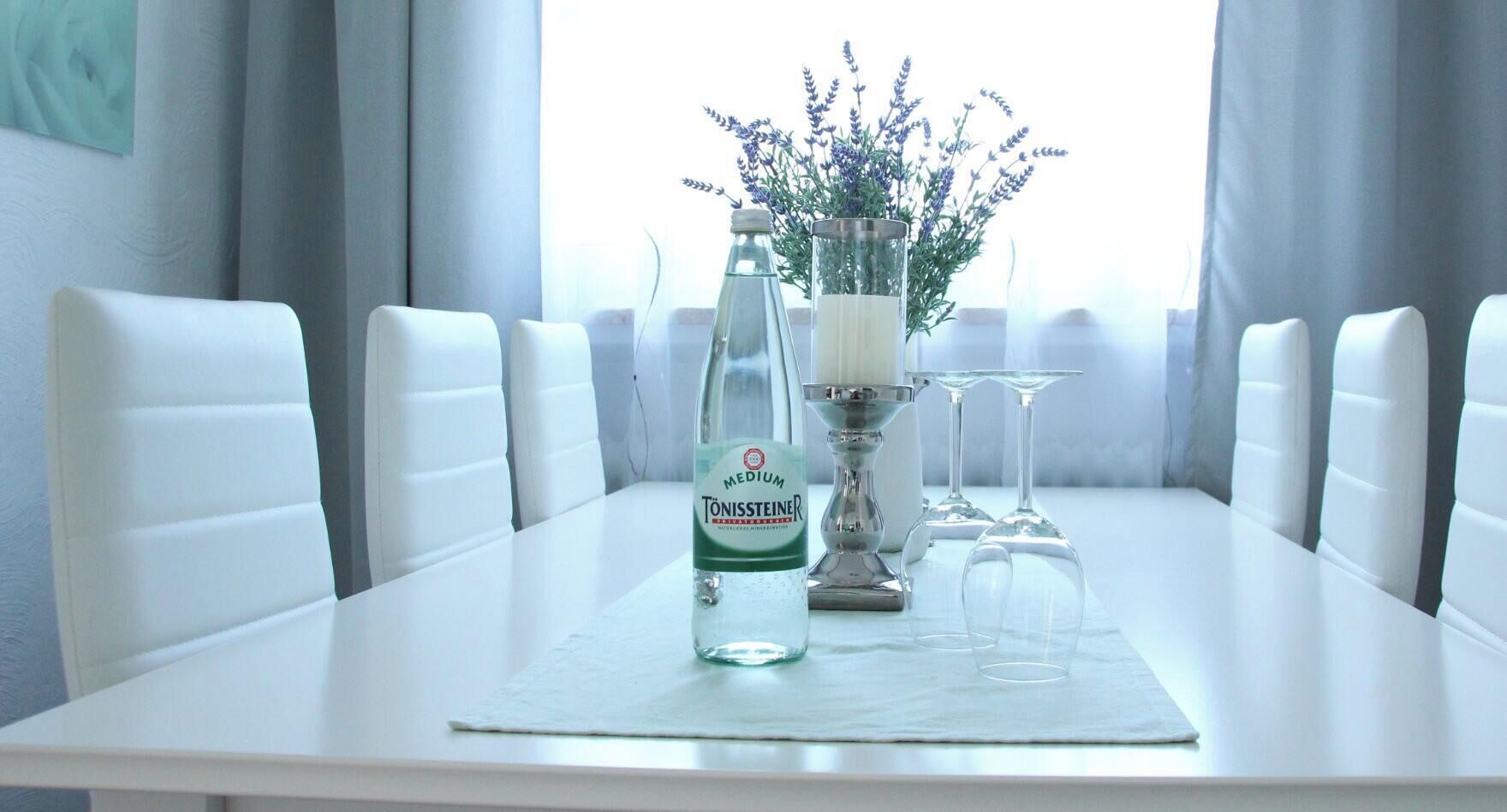 Et spisebord med hvide stole, en flaske tonic, en glasvas med lavendel og en sølvlysestage.