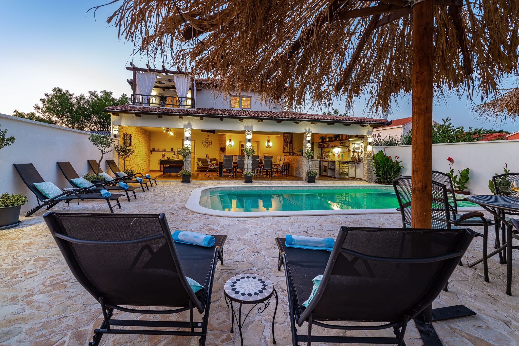 A poolside area with lounge chairs and a dining table, a pool, and a building with a tiled roof and a balcony.