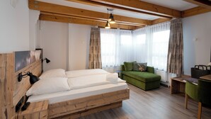 Una cama con estructura de madera y cabecera, un sofá verde, una mesa de madera y una ventana con cortinas de gasa.