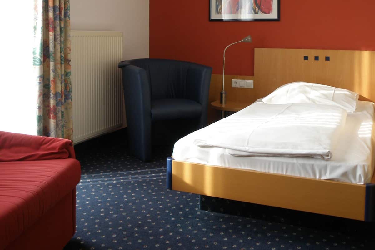 Habitació d'hotel amb un llit, un sofà vermell, un butac armchair blau, un quadre a la paret i una finestra amb cortines.