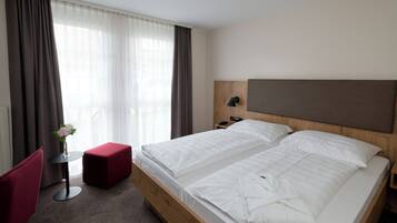 Ein Hotelzimmer mit einem Doppelbett, einem kleinen Tisch mit Blumenvase, einem roten Sessel und einem hölzernen Kopfteil.