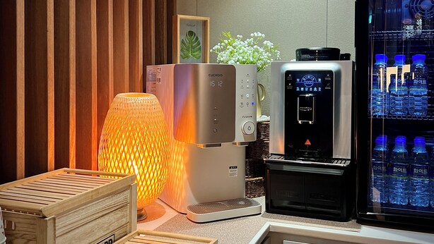 Een moderne keuken met een koffieapparaat, een waterdispenser en een houten bijzettafeltje met een lamp.