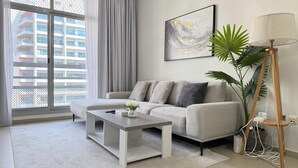 A modern living room with a grey sofa, a white coffee table, a floor lamp, and a large window with a view of a building. - Luxury Oasis Apartments in Barsha Heights (Dubai)