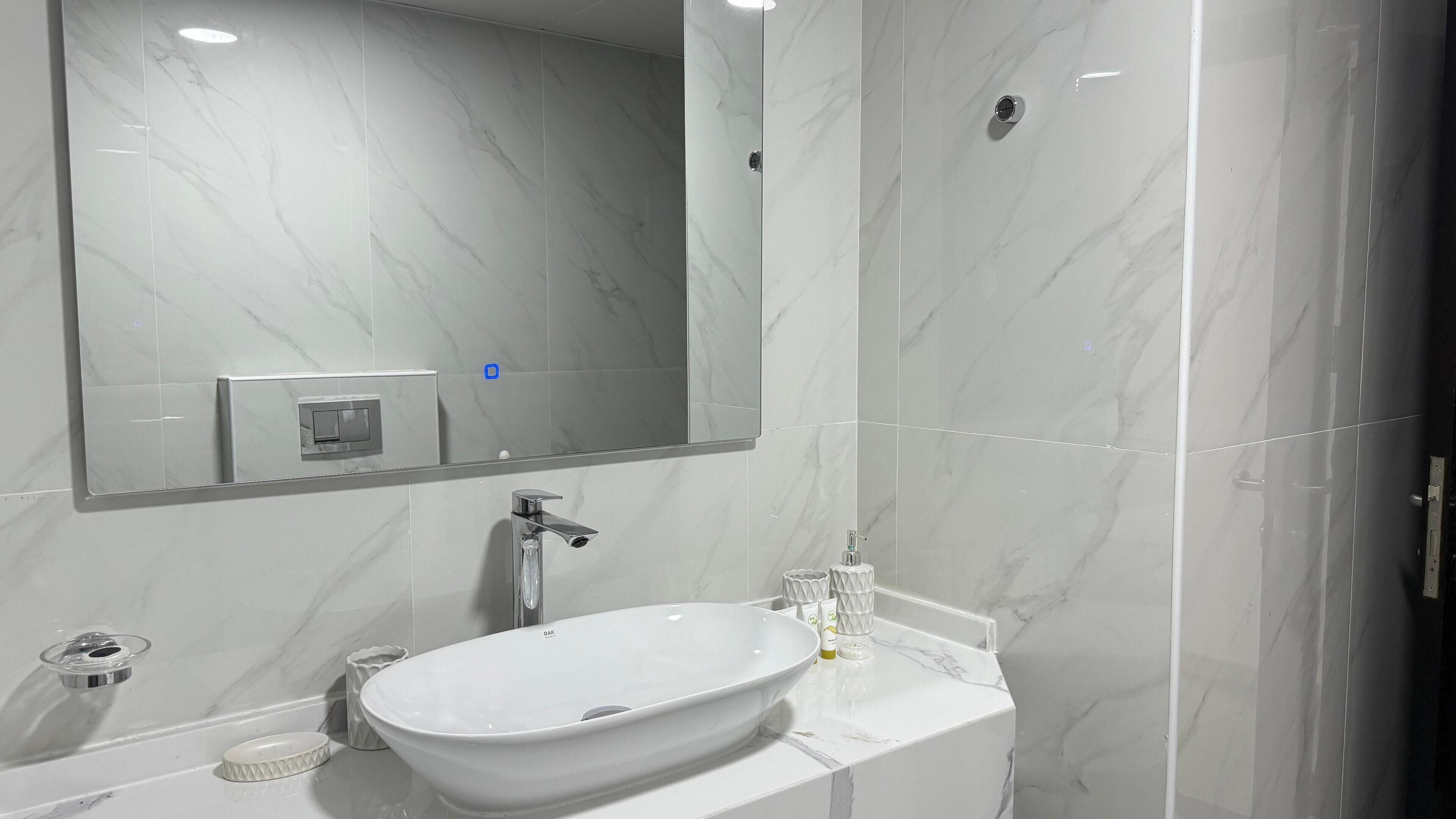 Une salle de bain avec un grand miroir, un lavabo blanc et un mur recouvert de carreaux de marbre.