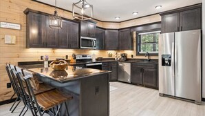 A modern kitchen with dark cabinetry, a central island with stools, stainless steel appliances, and a hanging light fixture. - Villa De Luxe by Beyond Expectations: Cabin w/ Indoor Pool, Hot Tub & Game Room (Pigeon Forge)