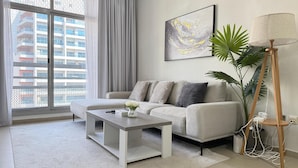 Un salon moderne avec un canapé gris, une table basse blanche, un lampadaire et une grande fenêtre donnant sur un immeuble.