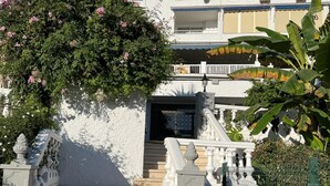 Un bâtiment à l’extérieur blanc, un escalier en béton menant à une porte sombre, et une clôture métallique ornée de verdure.