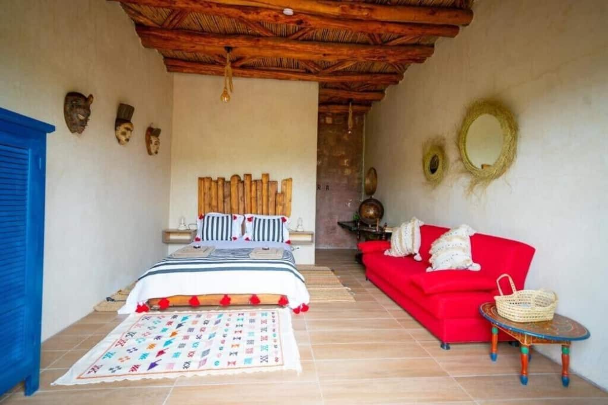 A bedroom with a wooden ceiling, a bed with striped bedding, a red sofa, and a small table with a basket.