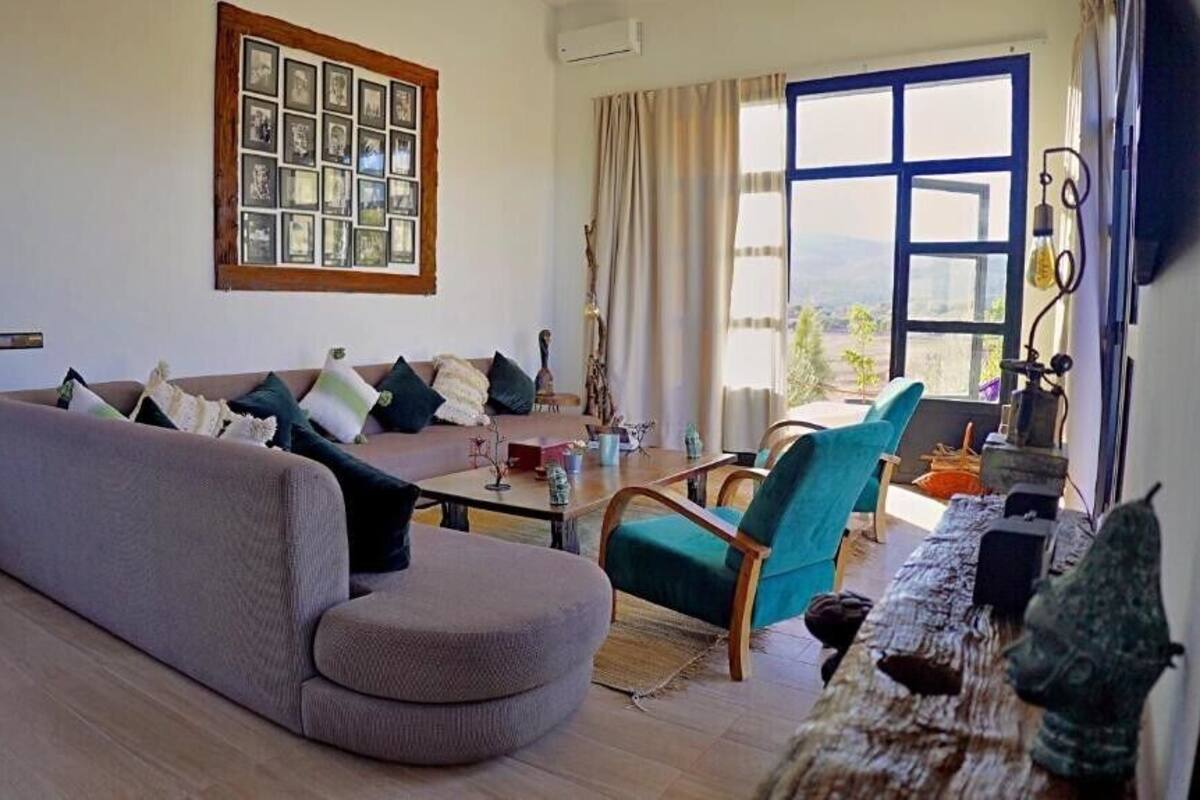 A living room with a sofa, armchairs, a coffee table, and a dining table. There is a large window with a view of the outdoors.