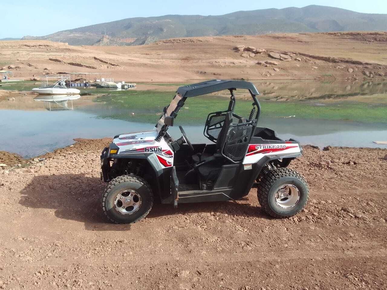 A utility vehicle parked on a dirt path near a body of water with a hilly landscape in the background.