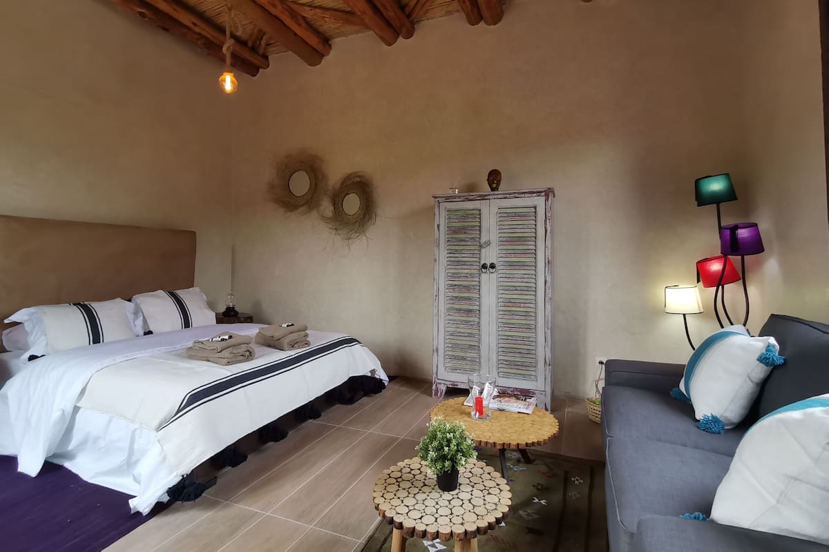 A bedroom with a bed, a small round table with a plant, a sofa, a wardrobe, and a wall-mounted lamp.