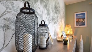 A room with two black wireframe lanterns, a lit table lamp, a framed picture, and a bottle on a wooden table. - Maison agréable près Etretat (Beaurepaire)
