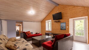 A cozy cabin-style room with a wooden ceiling, a flat-screen TV mounted above a sofa, a dining table, and a door leading to an outdoor balcony. - Holiday Home 'À Isturits Avec Piscine' with Shared Pool, Private Terrace and Wi-Fi (Isturits)