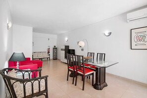 A modern living room with a dining area, a red sofa, a glass table, and a kitchen in the background. - Gabriel 6 H Apartment with Shared Pool, Balcony and Air Conditioning (Armação de Pêra)