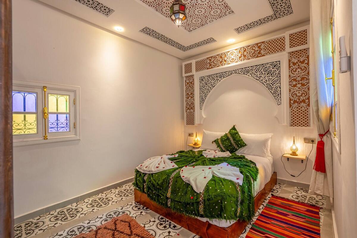 A bedroom with a bed, a nightstand, a wall-mounted lamp, and a window with decorative glass.