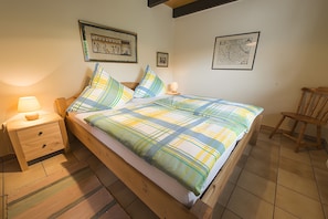 A neatly made bed with a plaid bedspread, a wooden nightstand with a lamp, and a chair in a room with tiled flooring. - Holiday Home 'Ferienhaus Schober' with Private Terrace, Garden and Wi-Fi (Cuxhaven)