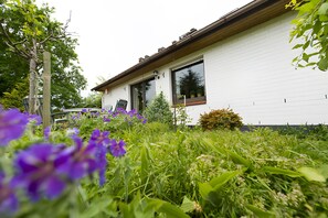 Une maison blanche avec un toit en bois, de grandes fenêtres et un jardin où poussent des fleurs violettes.