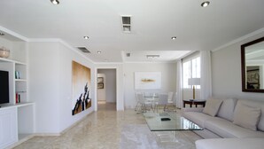 Amplia sala de estar con un sofá grande, una mesa de centro de vidrio y una mesa de comedor con sillas. Hay un televisor a la izquierda y obras de arte en las paredes.