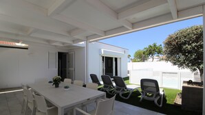 Un patio al aire libre con una mesa y sillas blancas, una zona cubierta y vistas a una casa y jardín.