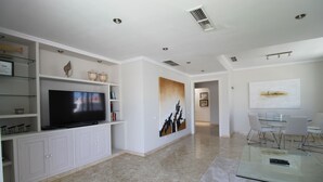 Una sala moderna con televisor de pantalla plana, muebles blancos, una mesa de centro de vidrio y un comedor con sillas.