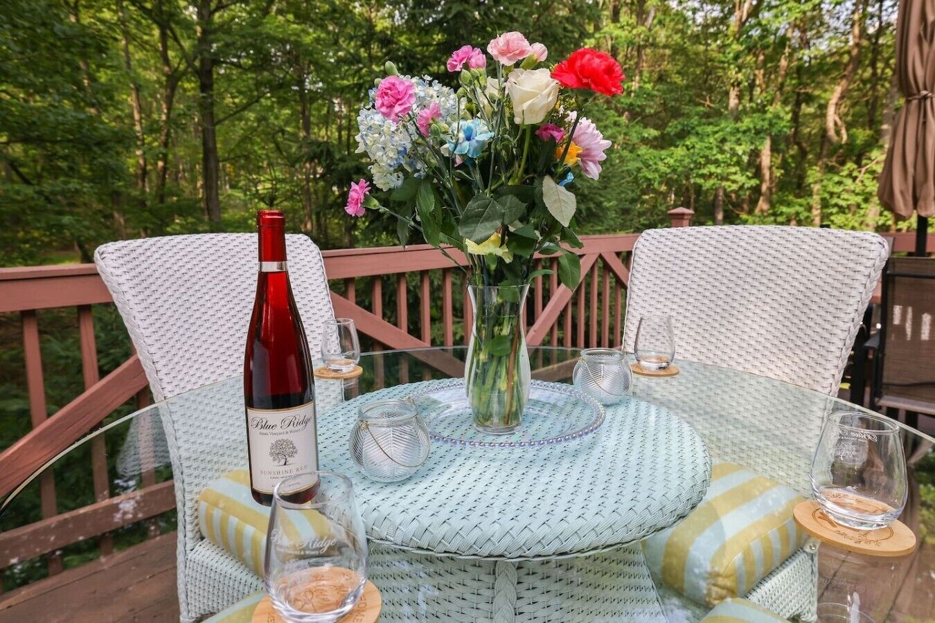 A round glass table with a white woven tablecloth, surrounded by wicker chairs with cushions. A vase with flowers and a bottle of wine are on the table.