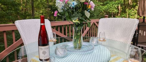 A round glass table with a white woven tablecloth, surrounded by wicker chairs with cushions. A vase with flowers and a bottle of wine are on the table.