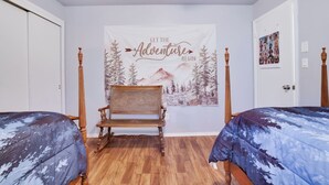 Ein Schlafzimmer mit zwei Betten, einem Holzstuhl, einer Wandteppich mit Berglandschaft und einer Tür mit Fotocollage.