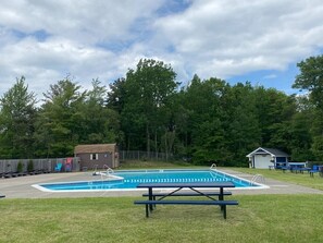 A swimming pool with a picnic table in the foreground, surrounded by trees and a small building in the background.