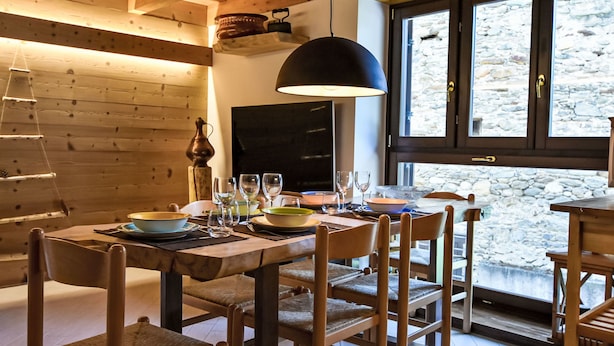 A wooden dining table with chairs, a hanging light fixture, and a stone wall with a window. - Ai Tini by Interhome (Campodolcino)