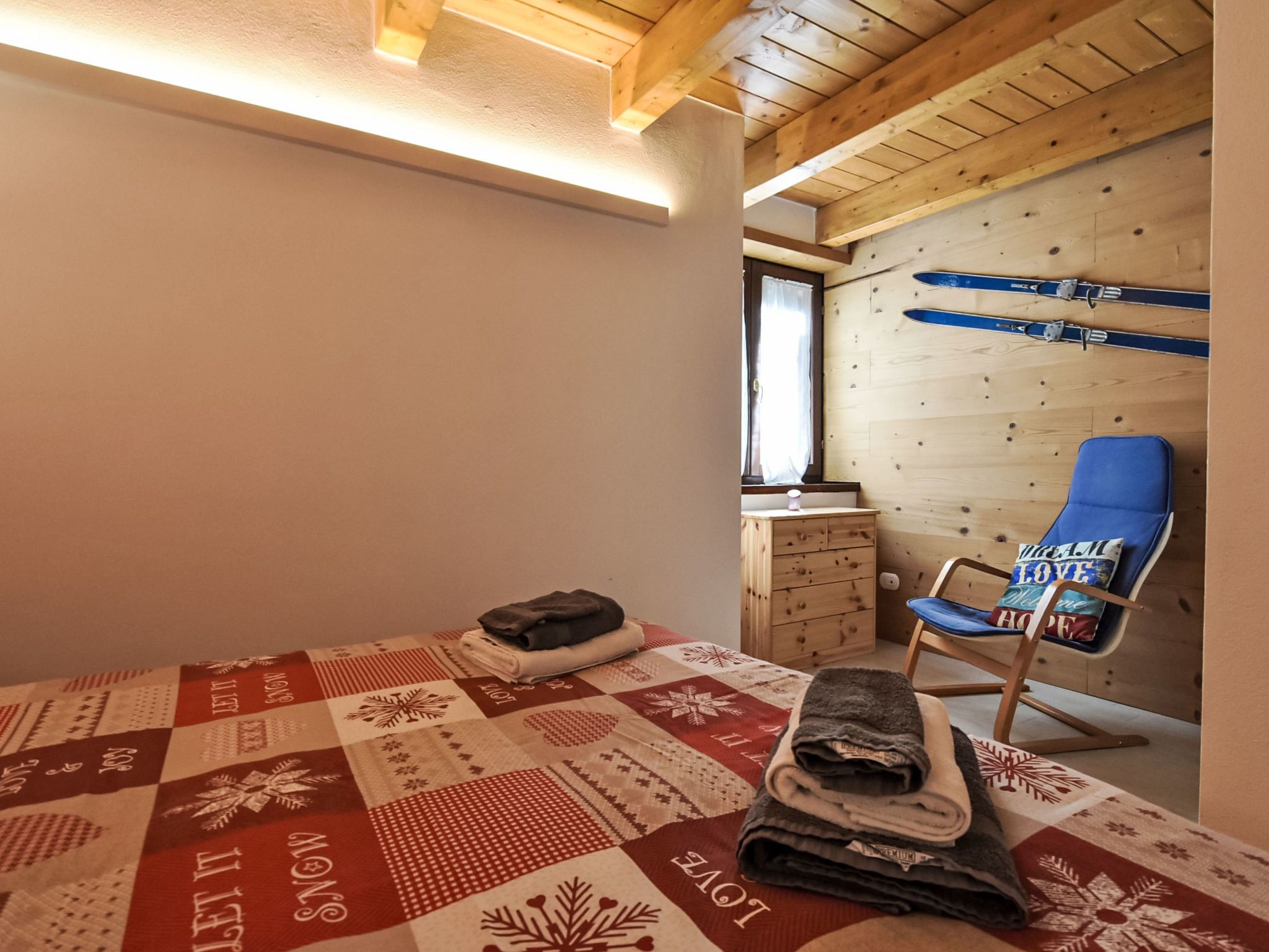 A cozy room with a wooden ceiling, a blue chair with a cushion, a quilted bedspread, and a wooden dresser.