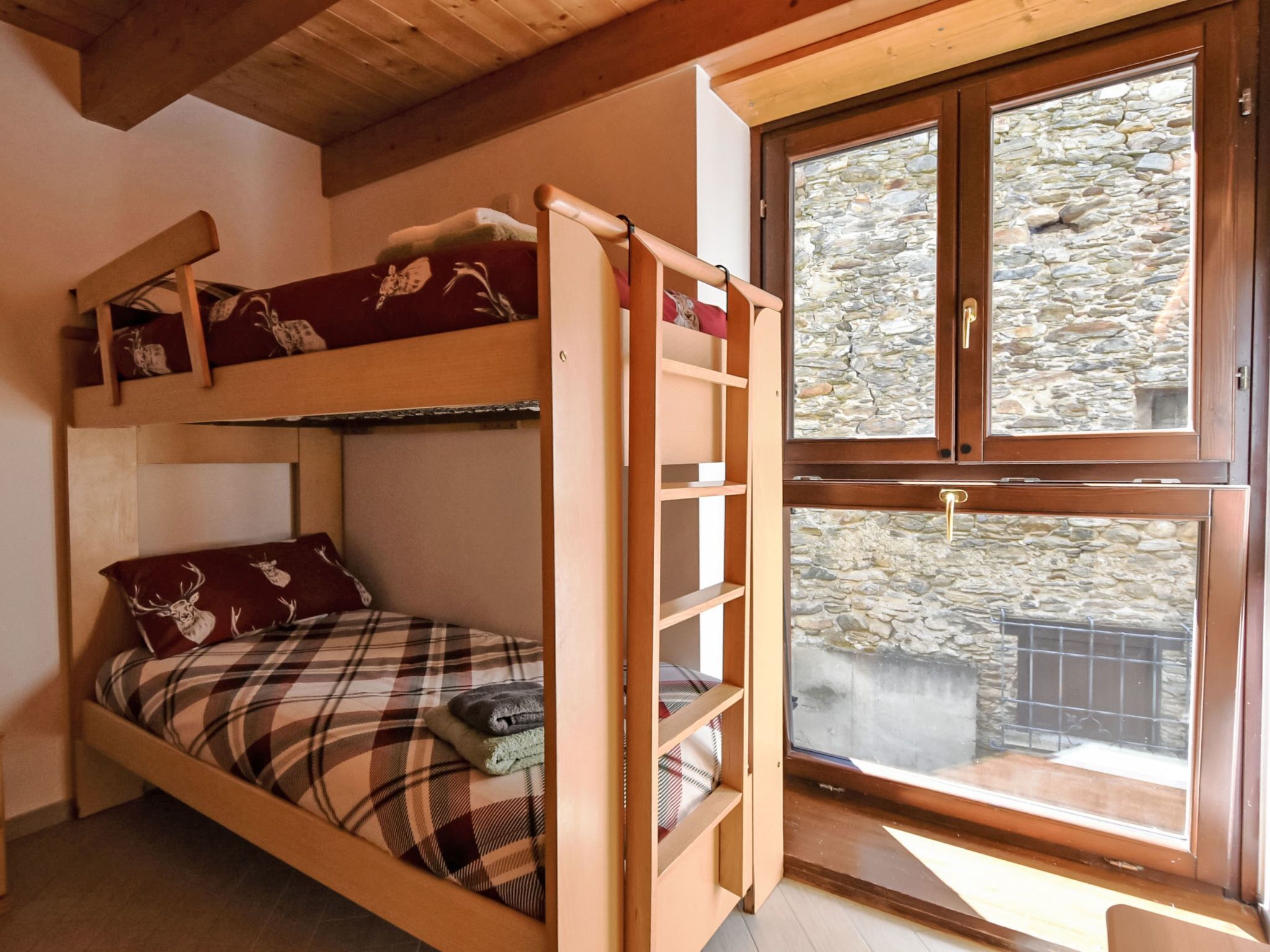 A bunk bed room with a wooden ladder, a plaid bedspread, and a window with a stone wall view.