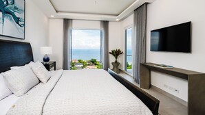 A modern hotel room with a large bed, a flat-screen TV, a ceiling fan, a window with a view of the sea, and a small desk with a lamp.