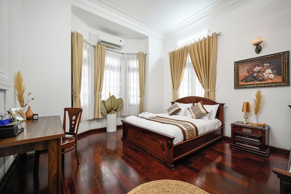 A bedroom with a wooden bed, beige curtains, a nightstand with a lamp, and a painting on the wall.