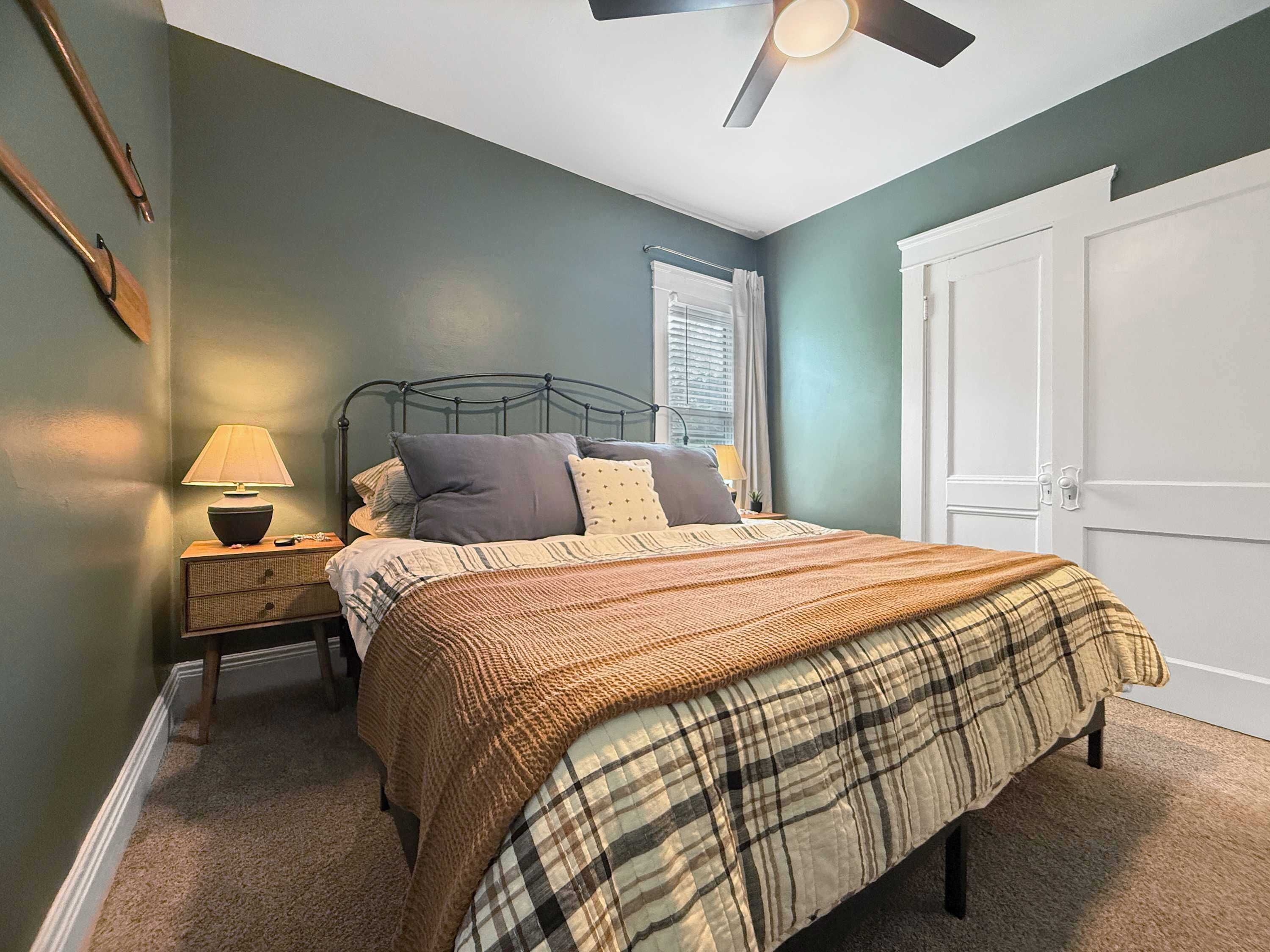 A bedroom with a bed, a nightstand, a lamp, a window with blinds, and a door.